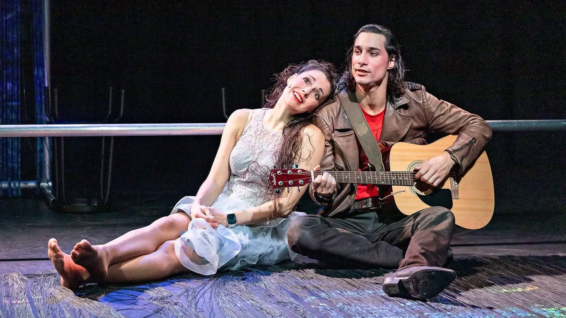 Ein Mann und eine Frau sitzen auf dem Boden einer Bühne und lehnen sich aneinander. Der Mann hält eine Gitarre in der Hand.
